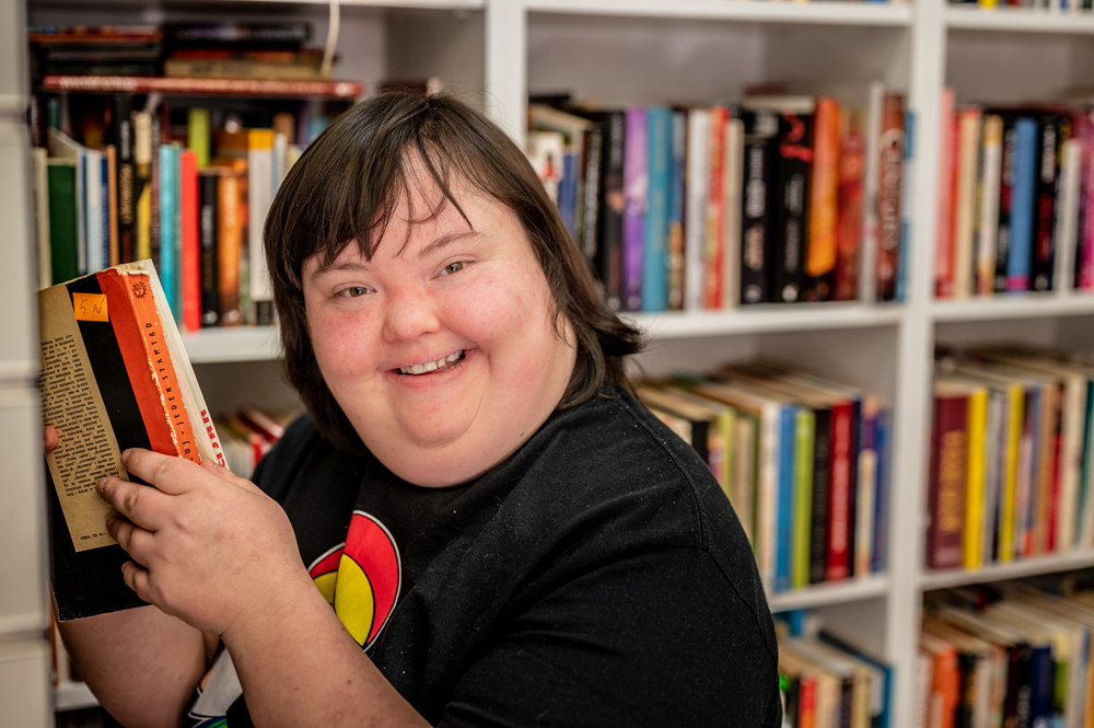 Zdjęcie przedstawia ciemnowłosą kobietę z zespołem Downa, uśmiechniętą, w czarnej koszulce. Trzyma dwie książki. Stoi na tle regałów z książkami. Pracuje w sklepie charytatywnym.