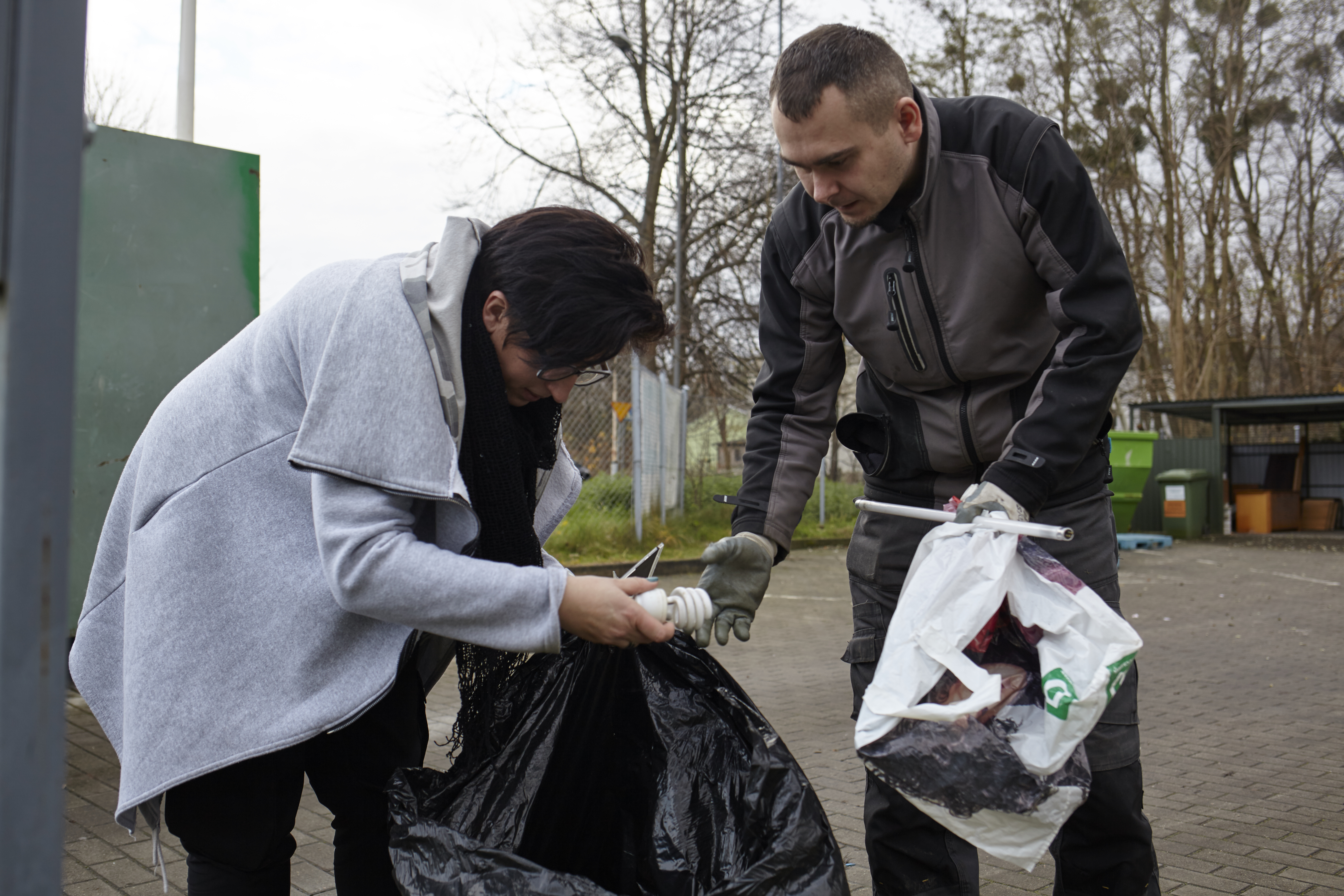 Na zdjęciu punkt zbiórki odpadów komunalnych. Na pierwszym planiu dwóch mieszkańców Szczecina - kobieta i mężczyzna. Kobieta trzyma w ręku otwarty worek foliowy, z którego wyjmuje żarówkę i podaje ją mężczyźnie.