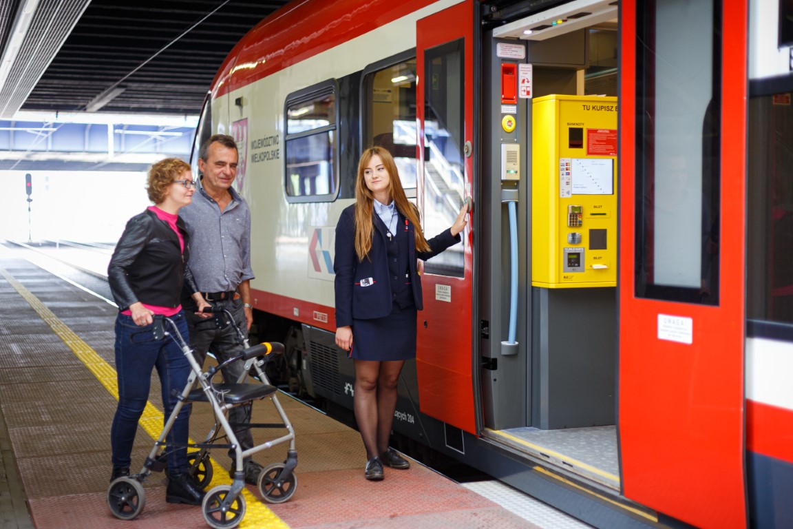 A woman with a walker close to a train with man and woman waiting for a ramp to be used.