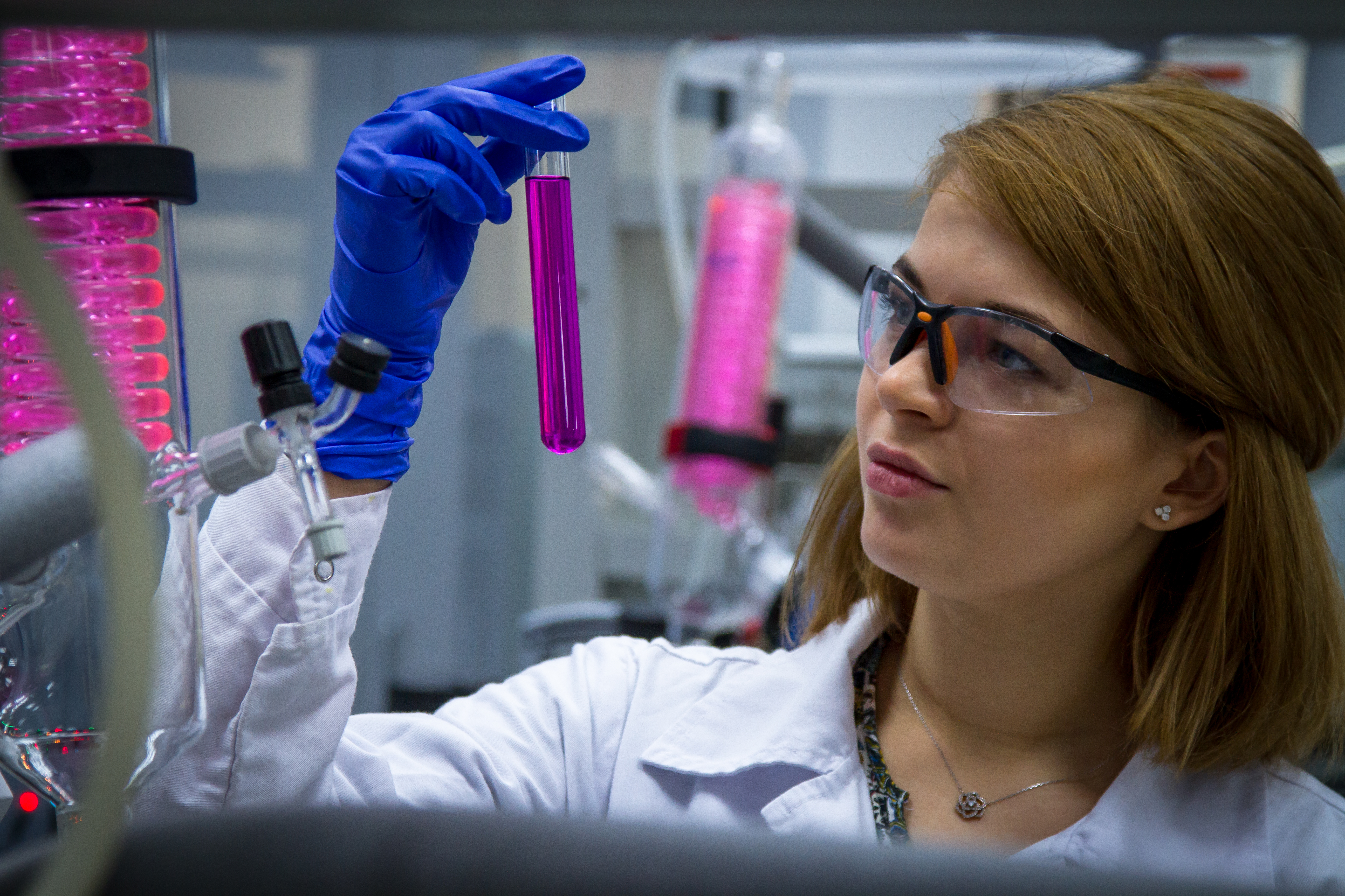 A female scientist wearing safety goggles holds a test tube with a pink substance in her hand.