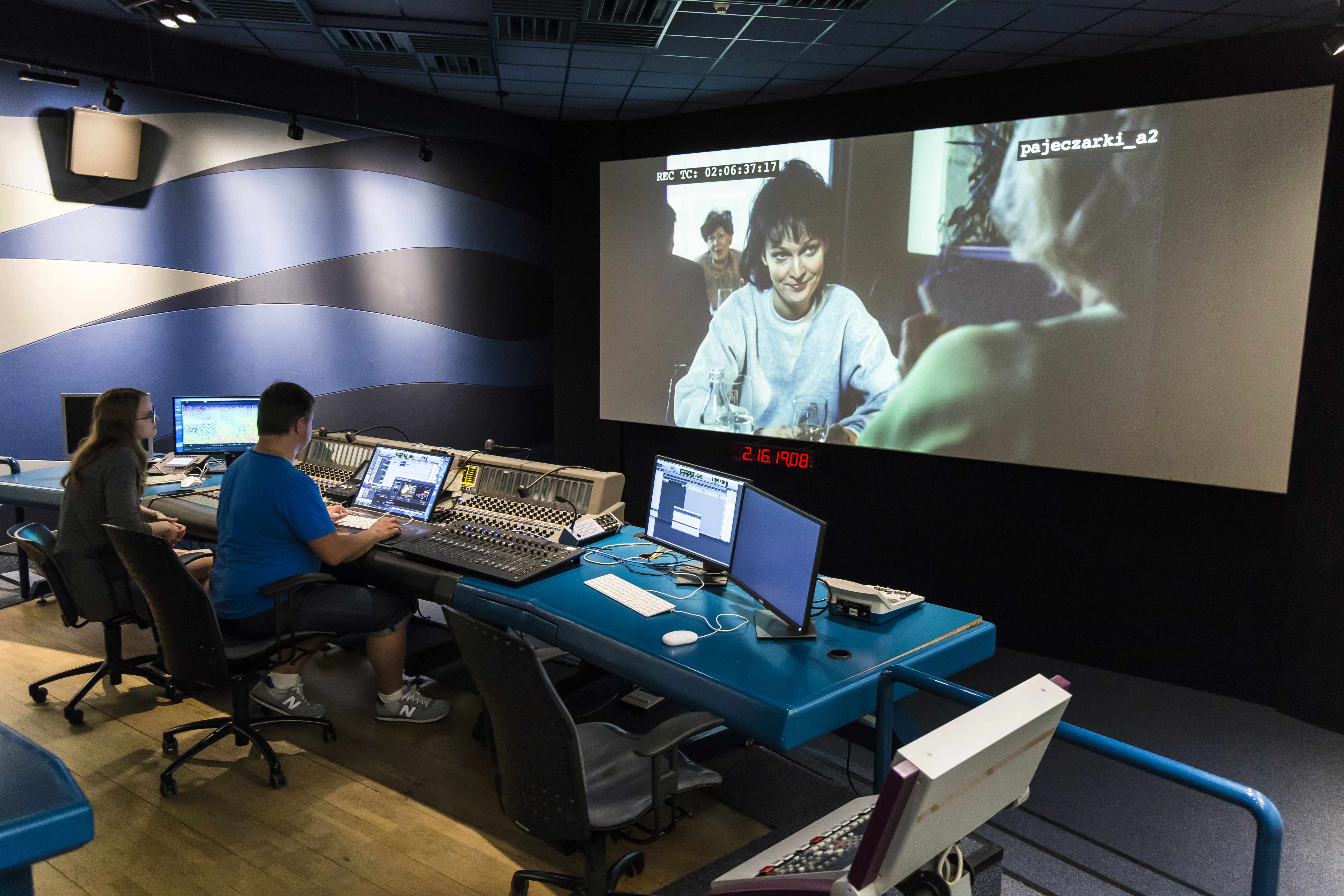 Zdjęcie przedstawia studio filmowe, z widocznym dużym ekranem kinowym oraz miejscem pracy reżysera.